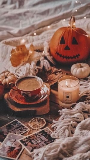 coffee cup next to lit candle carved pumpkin placed on wooden floor with white blankets aesthetic fall wallpaper