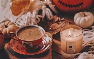 coffee cup next to lit candle carved pumpkin placed on wooden floor with white blankets aesthetic fall wallpaper