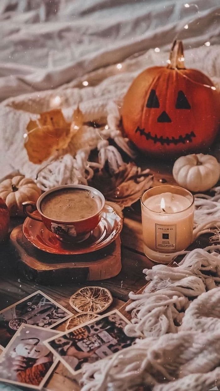 coffee cup next to lit candle carved pumpkin placed on wooden floor with white blankets aesthetic fall wallpaper